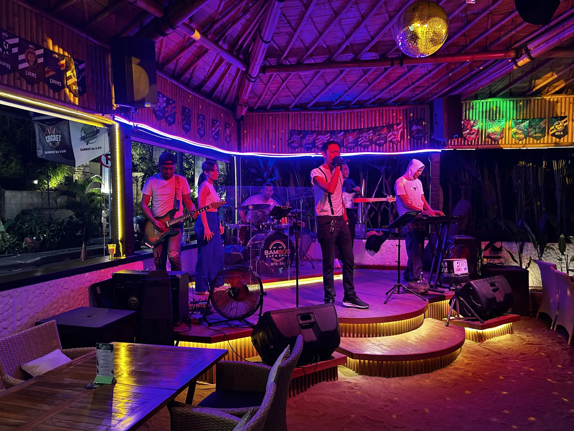 A live band on stage at Bamboo beneath the thatched bamboo roof, lit by neon strip lighting with a disco ball overhead